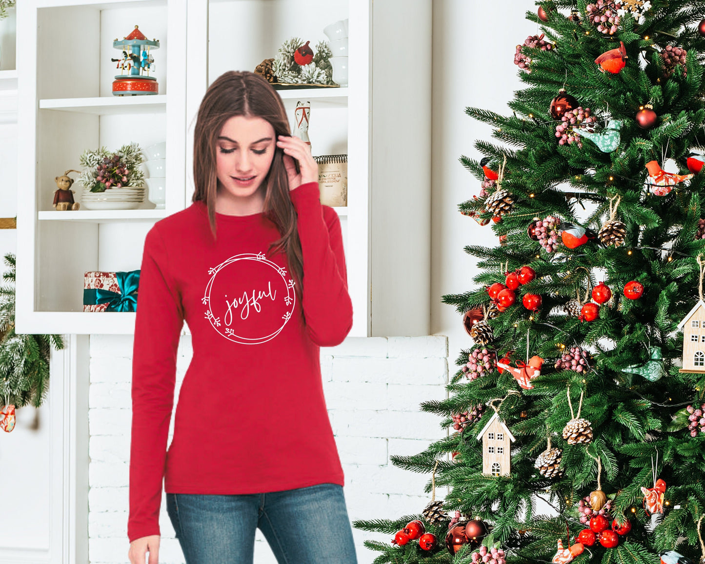 Woman standing next to a Christmas tree wearing a red long sleeve tee with the word "joyful" surrounded by a holiday wreath