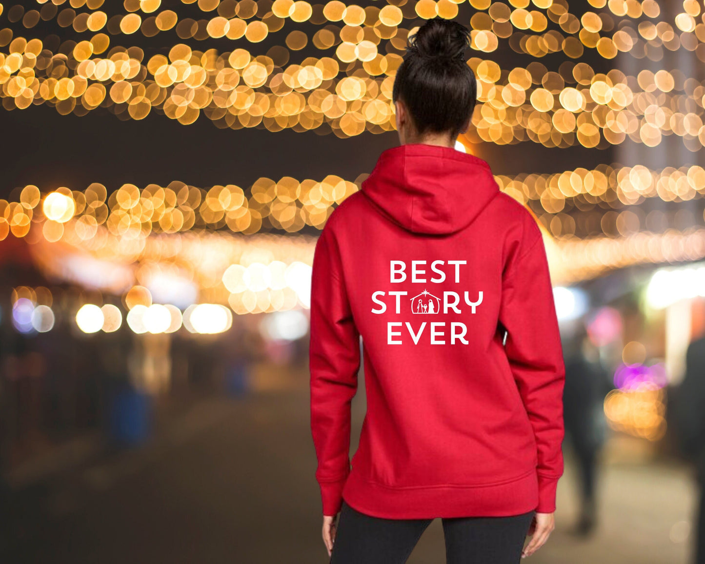 Woman standing in front of Christmas lights, wearing a red hoodie with the word "BEST STORY EVER" on the back. The "o" in "story" is replaced with a nativity scene.