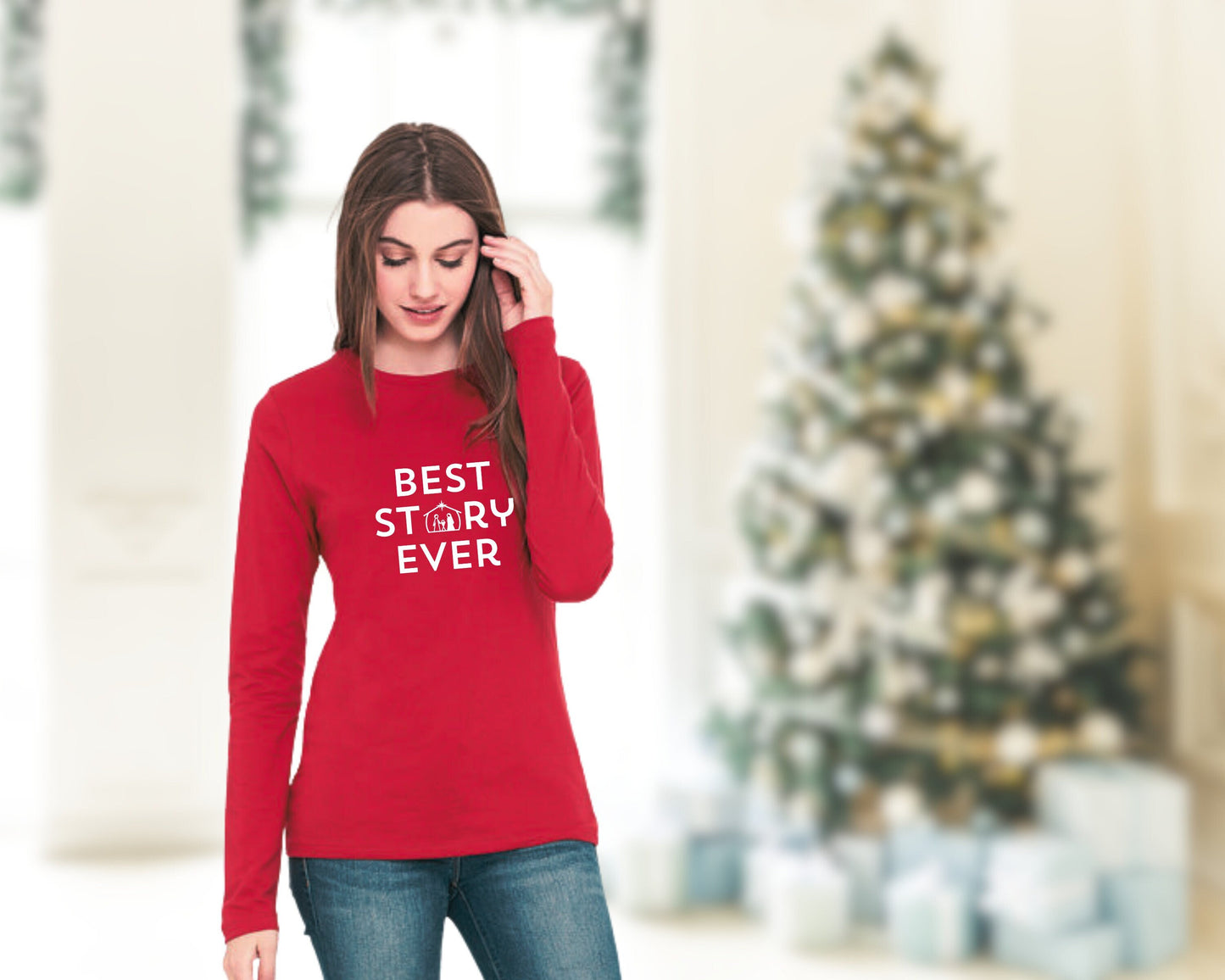 Woman standing in front a Christmas tree wearing a red long sleeve tee with the words "BEST STORY EVER". The nativity scene is incorporated into the text.