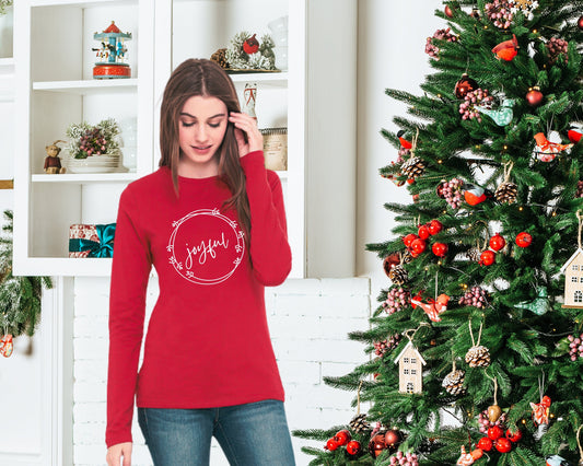 Woman standing next to a Christmas tree wearing a red long sleeve tee with the word &quot;joyful&quot; surrounded by a holiday wreath
